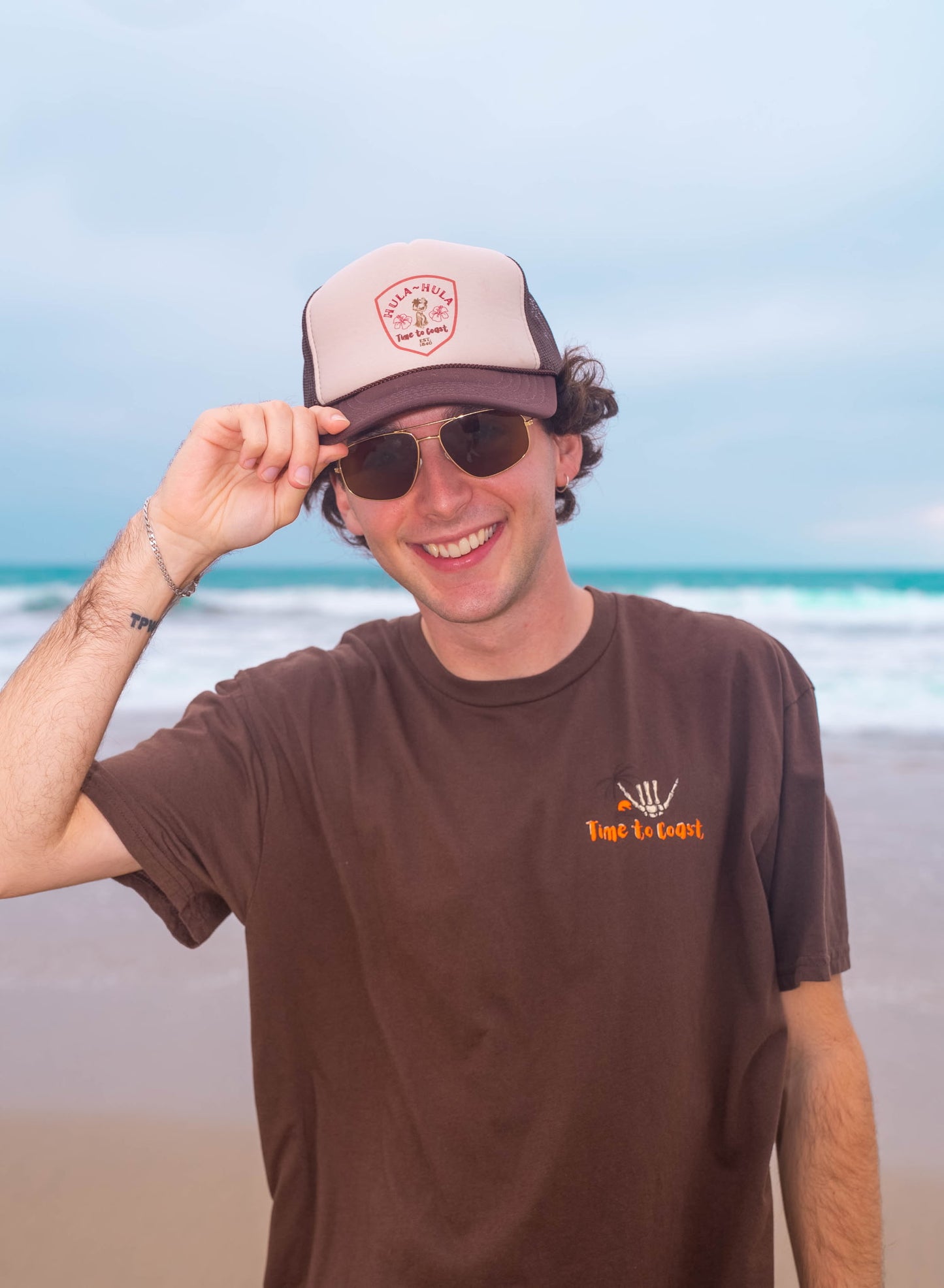 Man sunglasses on a beach wearing a foam high profile trucker hat and shaka tee. Graphic is printed with Time to Coast Hula Hula girl. Hat color is beige and brown with a brown mesh back.
