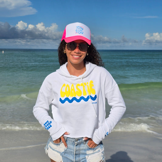 Woman standing on the beach by the ocean wearing sunglasses, denim shorts, Hippy Wave Foam High Profile hat and a Florida Fleece White Cropped Hoodie Graphic Top. Graphic is the Time to Coast Coastie Wave in yellow and blue. Sleeves have blue words of sandy toes & sun kissed nose.