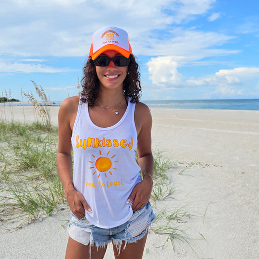 Woman in the sand dunes of the beach, wearing sunglasses, denim shorts and a white Time to Coast Palm Cloth Sunkissed Sun graphic tank top. Graphic color is orange.