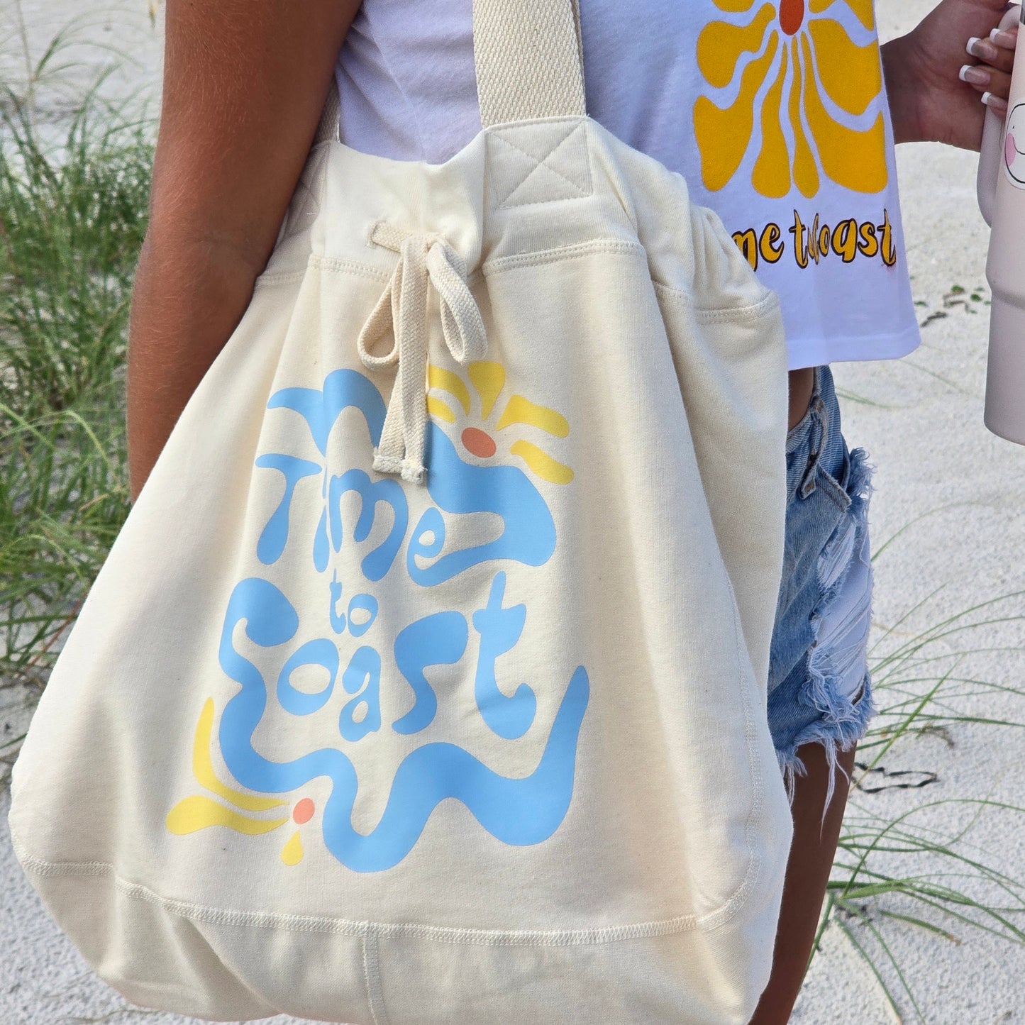 Model with hobo bag on her shoulder. The Florida Fleece Hobo Beach Bag, with drawstring closure. Creme colored with Time to Coast Hippy Wave Graphic in light blue and yellow.