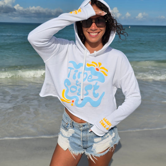 Woman standing on the beach by the ocean wearing sunglasses, denim shorts and a Florida Fleece White Cropped Hoodie Graphic Top. Graphic is the Time to Coast Hippy Wave in light blue and yellow. Both sleeves have yellow words of sandy toes & sunkissed nose.
