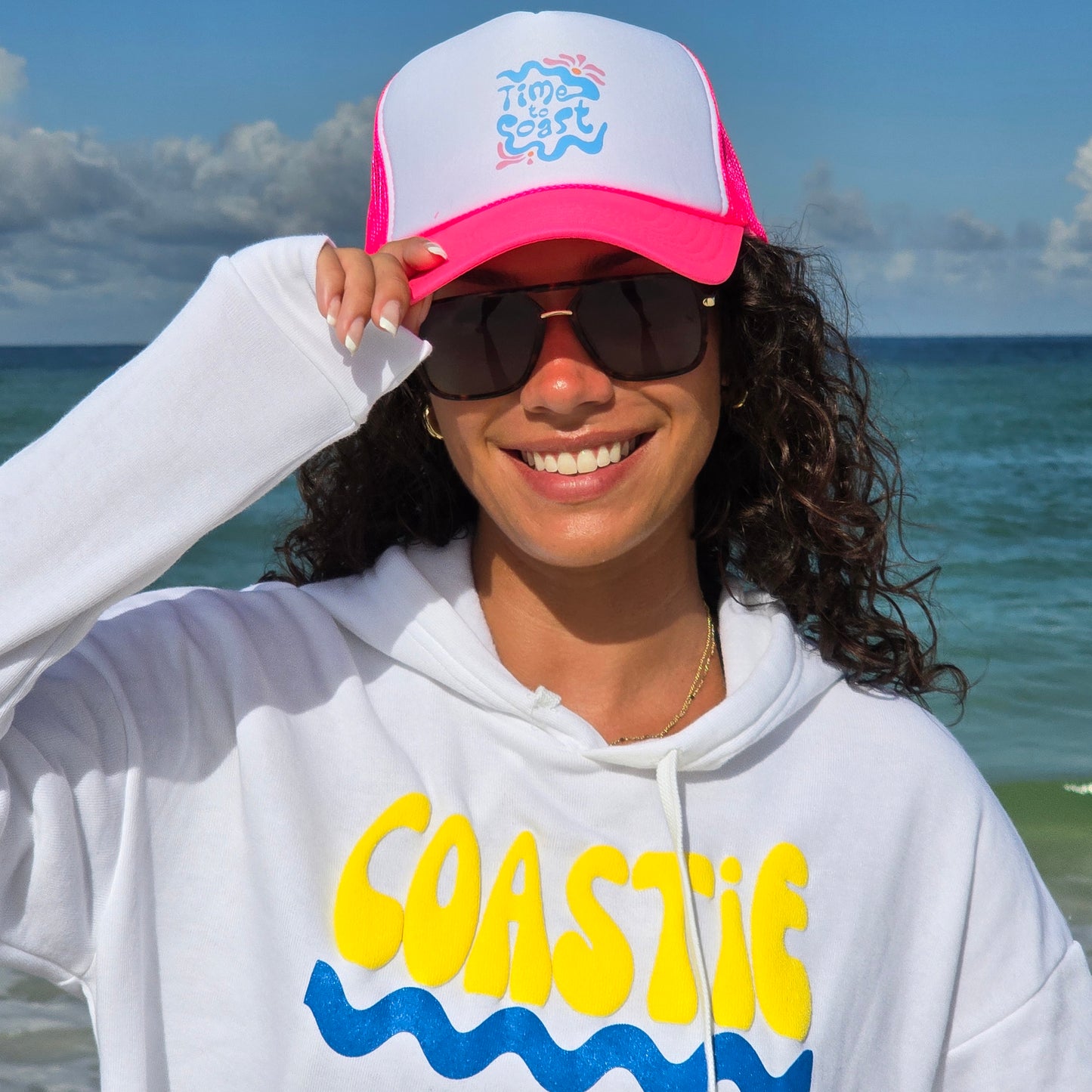 Woman wearing sunglasses, coastie crop florida fleece hoodie and a foam high profile trucker hat. Graphic is printed with Time to Coast Hippy Wave. Hat color is bright pink bill and mesh back with white front.