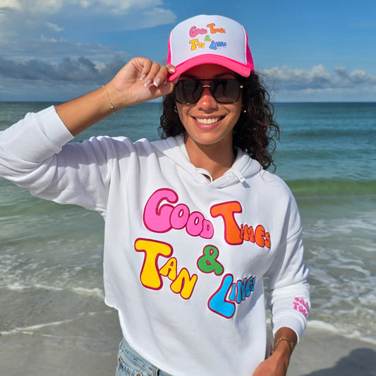 Woman standing on the beach by the ocean wearing sunglasses, denim shorts, Foam Good Times high profile hat and a Florida Fleece White Cropped Hoodie Graphic Top. Graphic is the Time to Coast Good Times & Tan Lines Slogan. Graphic is in multi colorful words. Sleeves have hot pink words of sandy toes & sun kissed nose.