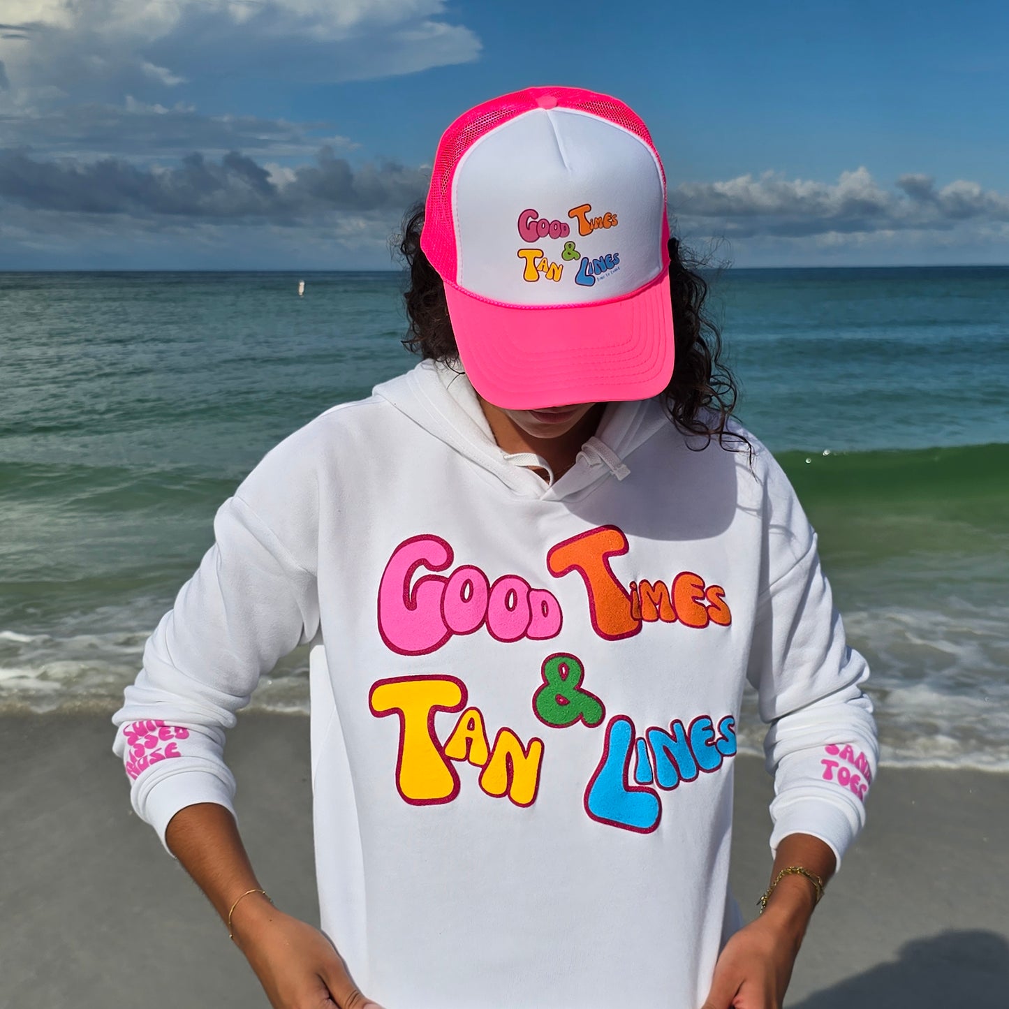 Woman on a beach wearing a foam high profile trucker hat. And a Crop Florida Fleece hoodie. Graphic is printed with Time to Coast Good Times and Tan Lines slogan. Hat color is bright pink bill and mesh back with white front.
