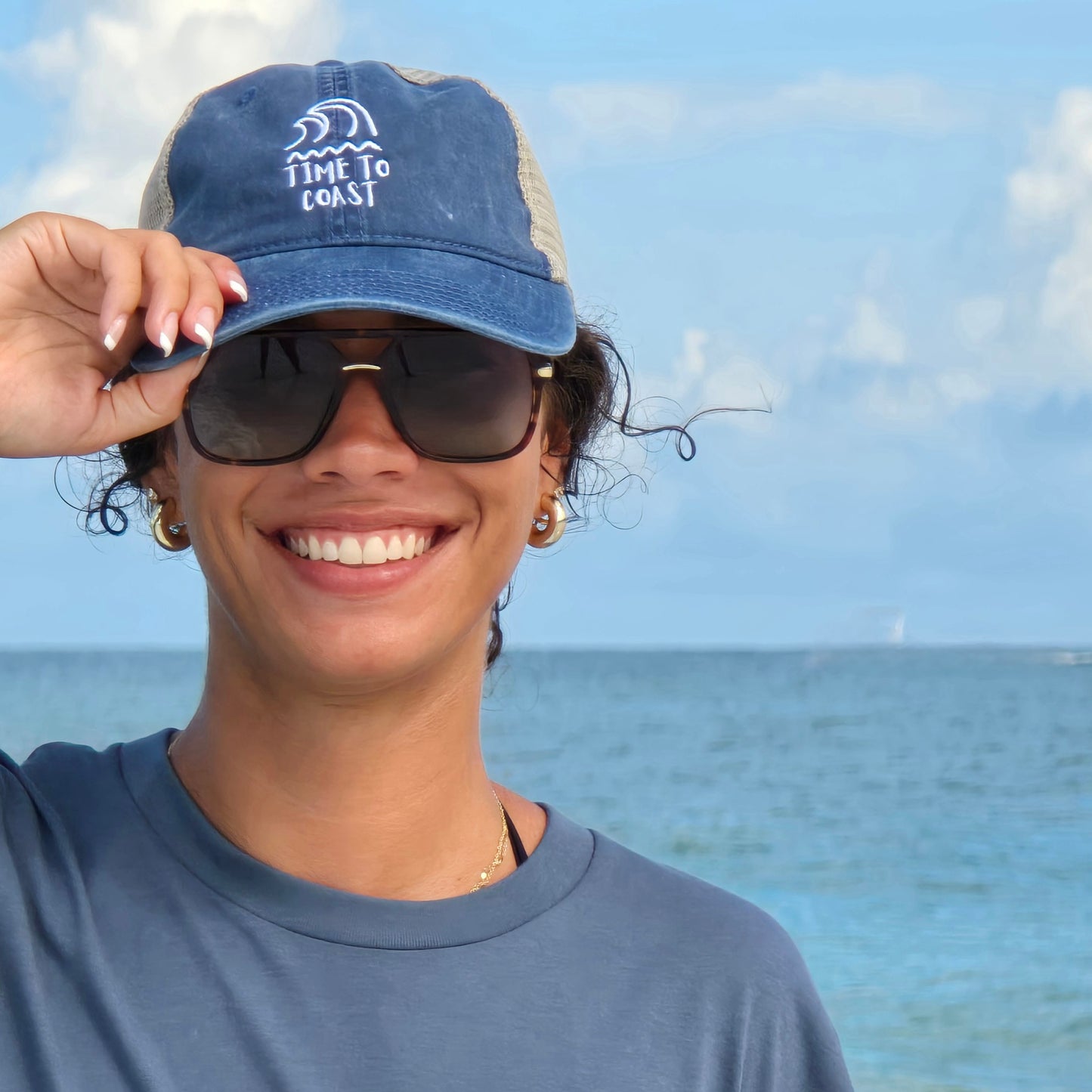 woman wearing sunglasses and a Time to Coast Swell wave twill low profile trucker hat. Hat is in a washed navy color with a tan mesh back. Graphic is an embroidery of surf swell and waves with time to coast in white.