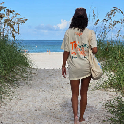 woman walking to the beach wearing a brown trucker hat. She is showing the back of the graphic tee unsex  graphic  t-shirt in organic cotton made in USA. Homegrown t-shirt shown with colorful back graphic of a Tiki Truck and Time to Coast Logo