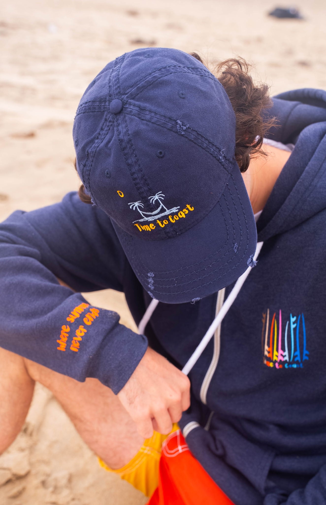 man sitting on the beach with his head down wearing the distressed dad hat in navy.  Embroidered graphic of the Time to Coast palm tree logo in light blue, yellow time to coast and sun