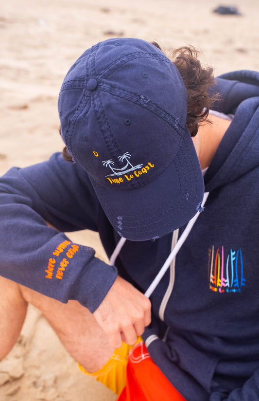 man sitting on the beach with his head down wearing the distressed dad hat in navy.  Embroidered graphic of the Time to Coast palm tree logo in light blue, yellow time to coast and sun