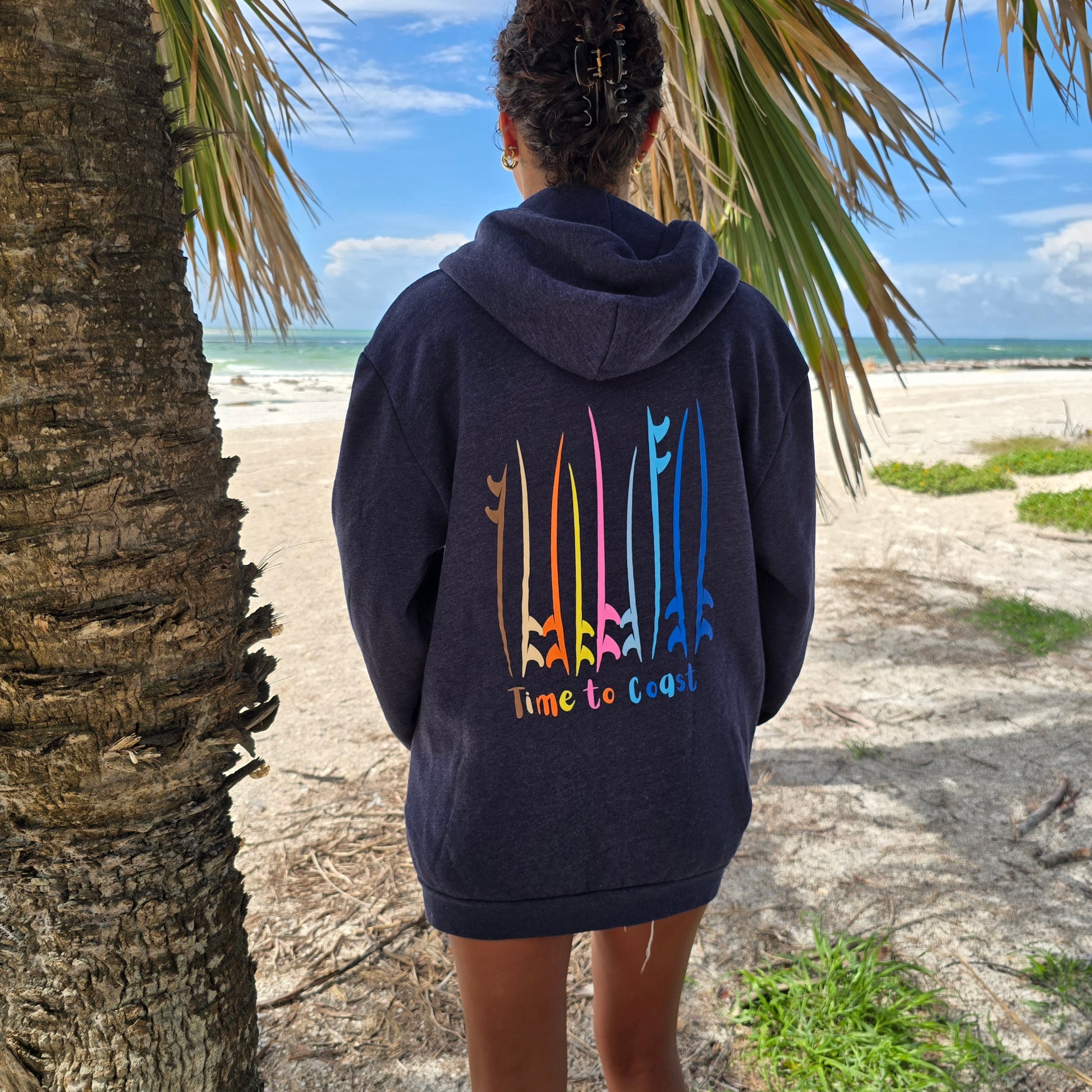 Woman on a beach looking out to the ocean, showing the back of the blue indigo Florida Fleece Zip Front Hoodie.  The graphic is the board meeting surf and sup boards in multi colors. Large graphic on back with small left chest graphic.  One sleeve has the words where summer never ends. Model is wearing a boyfriend fit.