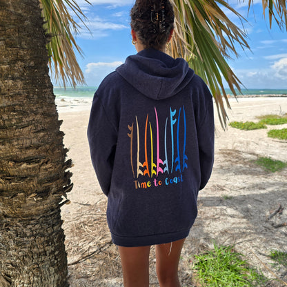 Woman on a beach looking out to the ocean, showing the back of the blue indigo Florida Fleece Zip Front Hoodie.  The graphic is the board meeting surf and sup boards in multi colors. Large graphic on back with small left chest graphic.  One sleeve has the words where summer never ends. Model is wearing a boyfriend fit.