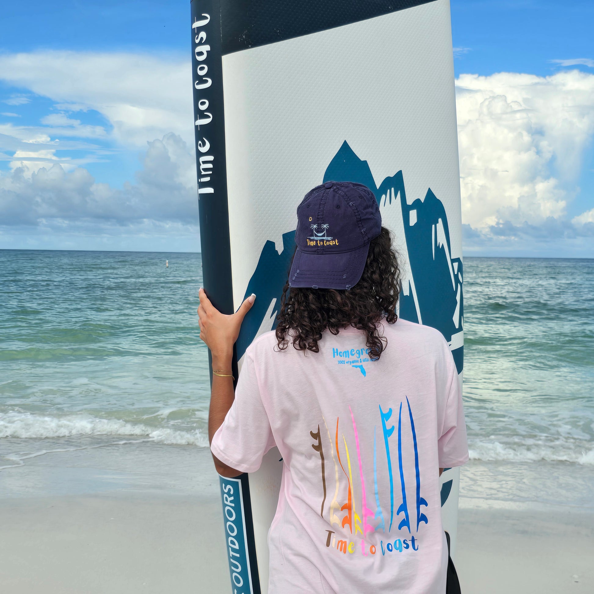 woman holding a sup board on the beach, wearing a navy distressed dad hat and a  graphic  t-shirt in organic cotton made in USA. Homegrown t-shirt with left chest and large back graphic of surf and sup boards. Board meeting graphic is surf or sup boards. Model is wearing a boyfriend fit size.