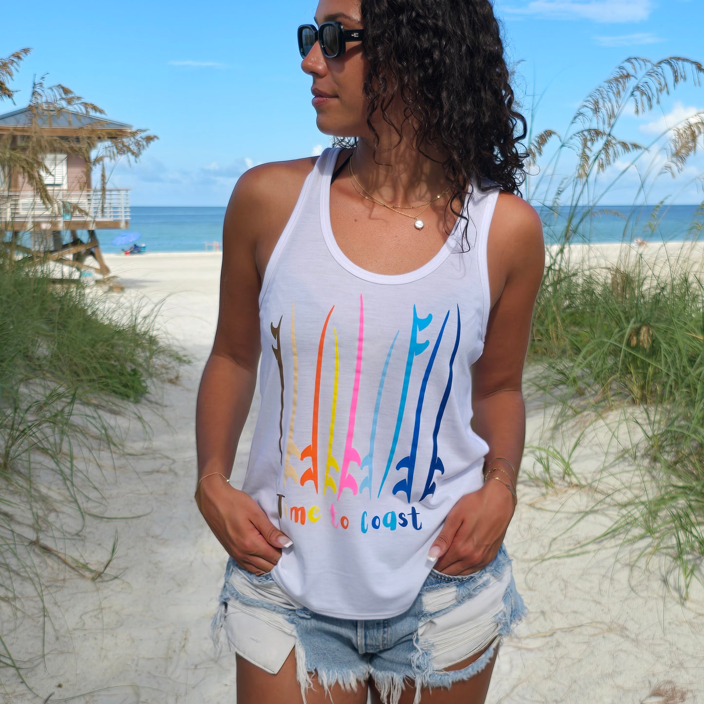 Woman in the sand dunes of the beach, wearing sunglasses, denim shorts and a white Time to Coast Palm Cloth Board meeting graphic tank top. Boards are surf and sup boards in bright colors.