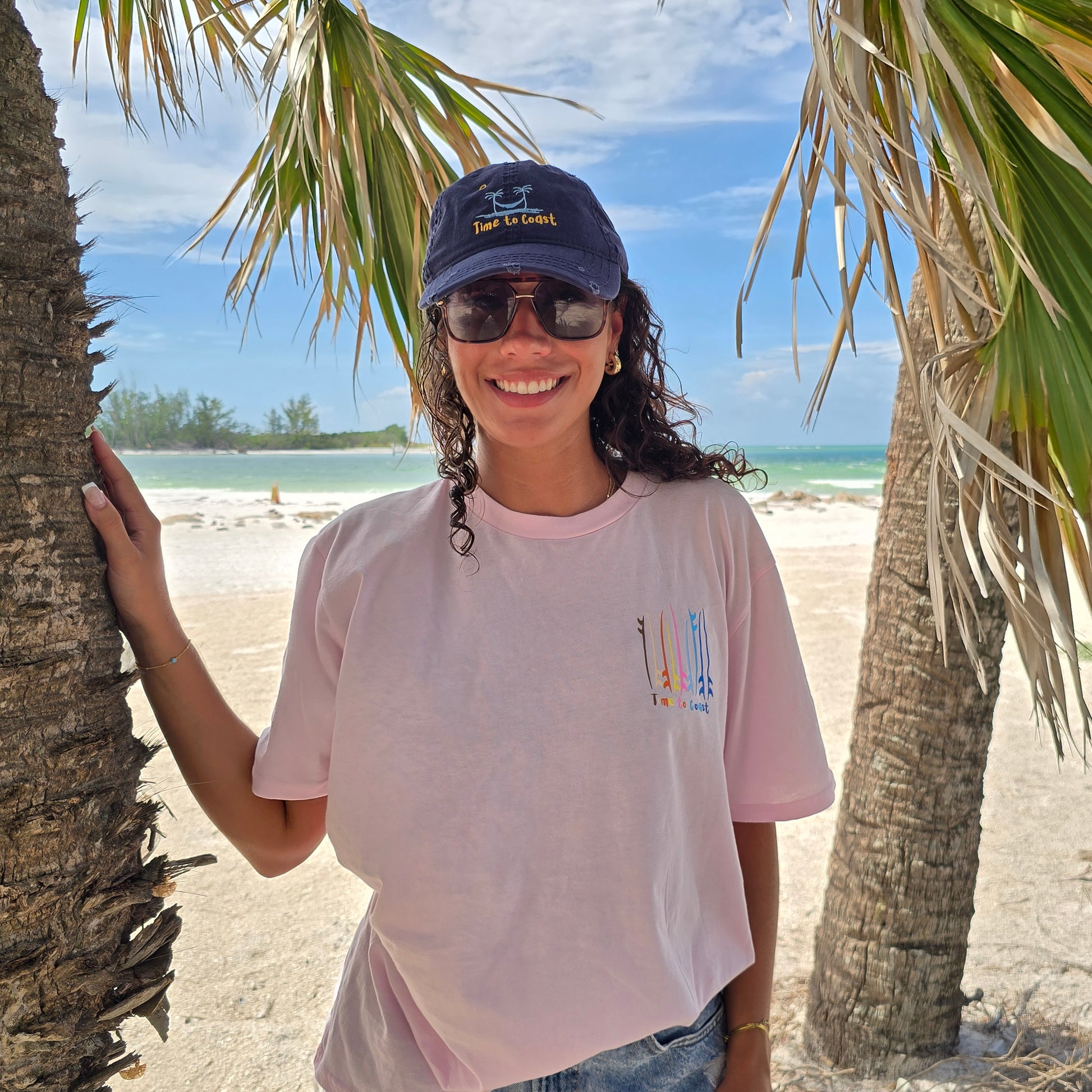 woman standing between palm trees on the beach wearing a navy distressed dad hat and a light pink   graphic  t-shirt in organic cotton made in USA. Homegrown t-shirt with left chest small  graphic of surf and sup boards.  Model is wearing a boyfriend fit size.