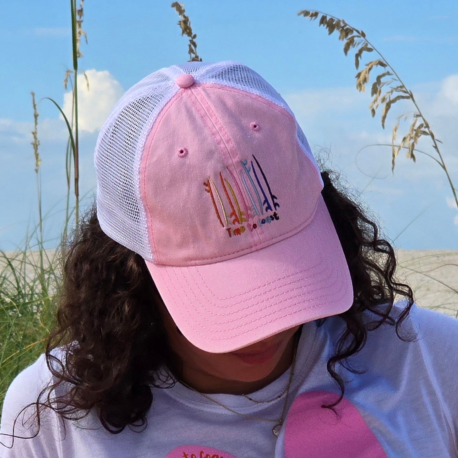 Woman on the sand dunes of the a beach wearing the Board Meeting embroidered Time to Coast washed pink twill low profile trucker hat with tan mesh back. Embroidered graphic is of surf and sup boards in multi colors.