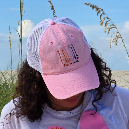 Woman on the sand dunes of the a beach wearing the Board Meeting embroidered Time to Coast washed pink twill low profile trucker hat with tan mesh back. Embroidered graphic is of surf and sup boards in multi colors.