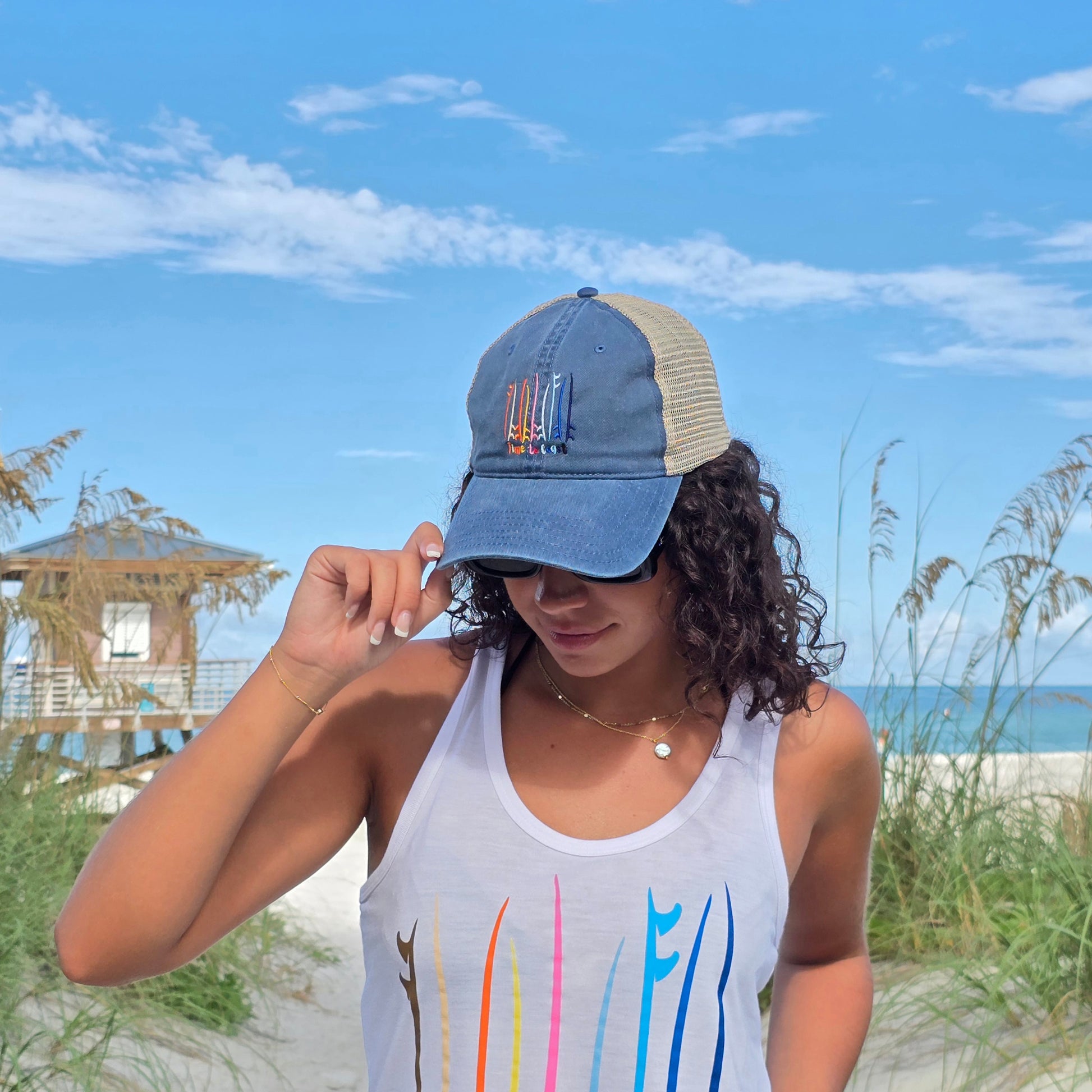 Woman on the sand dunes of the a beach wearing the Board Meeting embroidered Time to Coast washed navy twill low profile trucker hat with tan mesh back. Embroidered graphic is of surf and sup boards in multi colors.