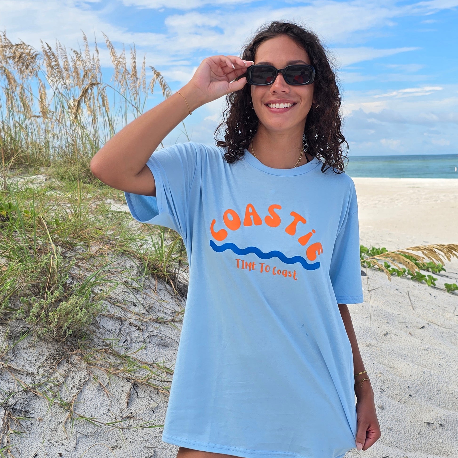 woman in the beach sand dunes, wearing sunglasses and a light blue  graphic  t-shirt in organic cotton made in USA. Homegrown t-shirt with large word graphic, of Coastie in orange and blue waves at the bottom.  Small Time to Coast Logo.  Model is wear a boyfriend fit size. 