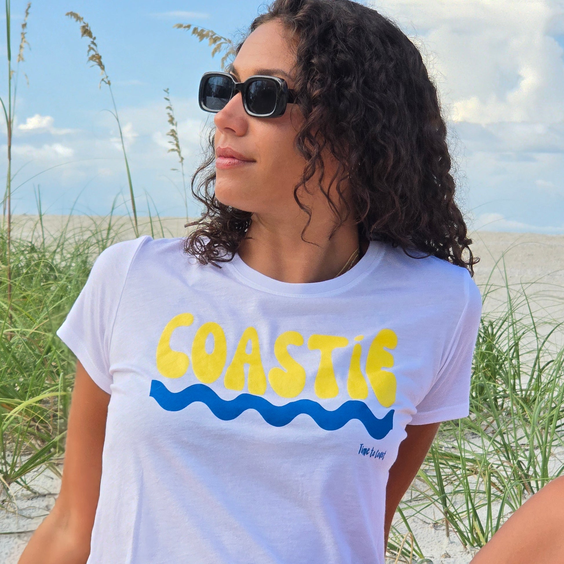 Woman sitting in the sand dunes of the beach, wearing sunglasses, and a white Time to Coast Palm Cloth Coastie crop Tee. Coastie graphic is yellow with a blue wave. 