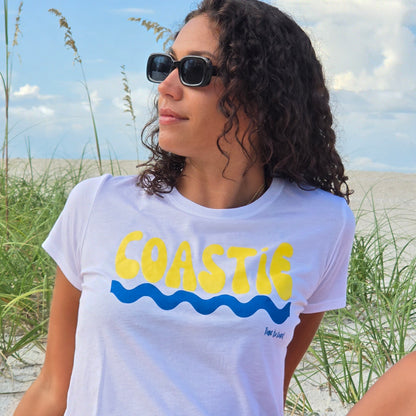 Woman sitting in the sand dunes of the beach, wearing sunglasses, and a white Time to Coast Palm Cloth Coastie crop Tee. Coastie graphic is yellow with a blue wave. 