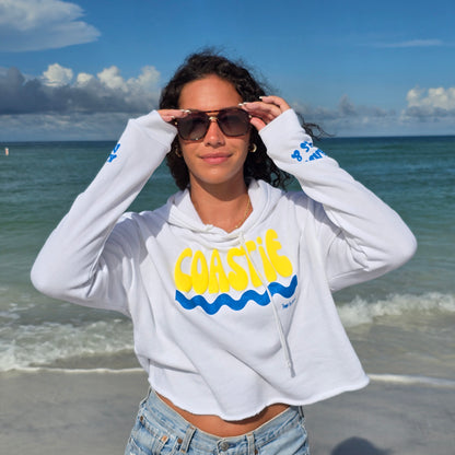 Woman standing on the beach by the ocean wearing sunglasses, denim shorts and a Florida Fleece White Cropped Hoodie Graphic Top.  Graphic is the Time to Coast Coastie Wave in yellow and blue.  Sleeves have blue words of sandy toes & sun kissed nose. 