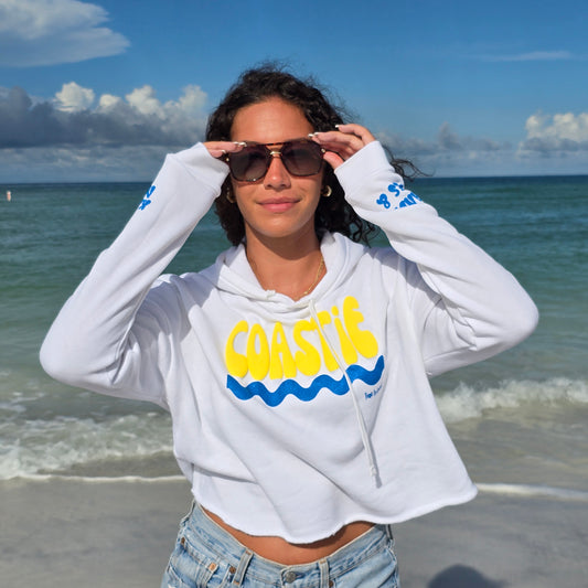 Woman standing on the beach by the ocean wearing sunglasses, denim shorts and a Florida Fleece White Cropped Hoodie Graphic Top.  Graphic is the Time to Coast Coastie Wave in yellow and blue.  Sleeves have blue words of sandy toes & sun kissed nose. 