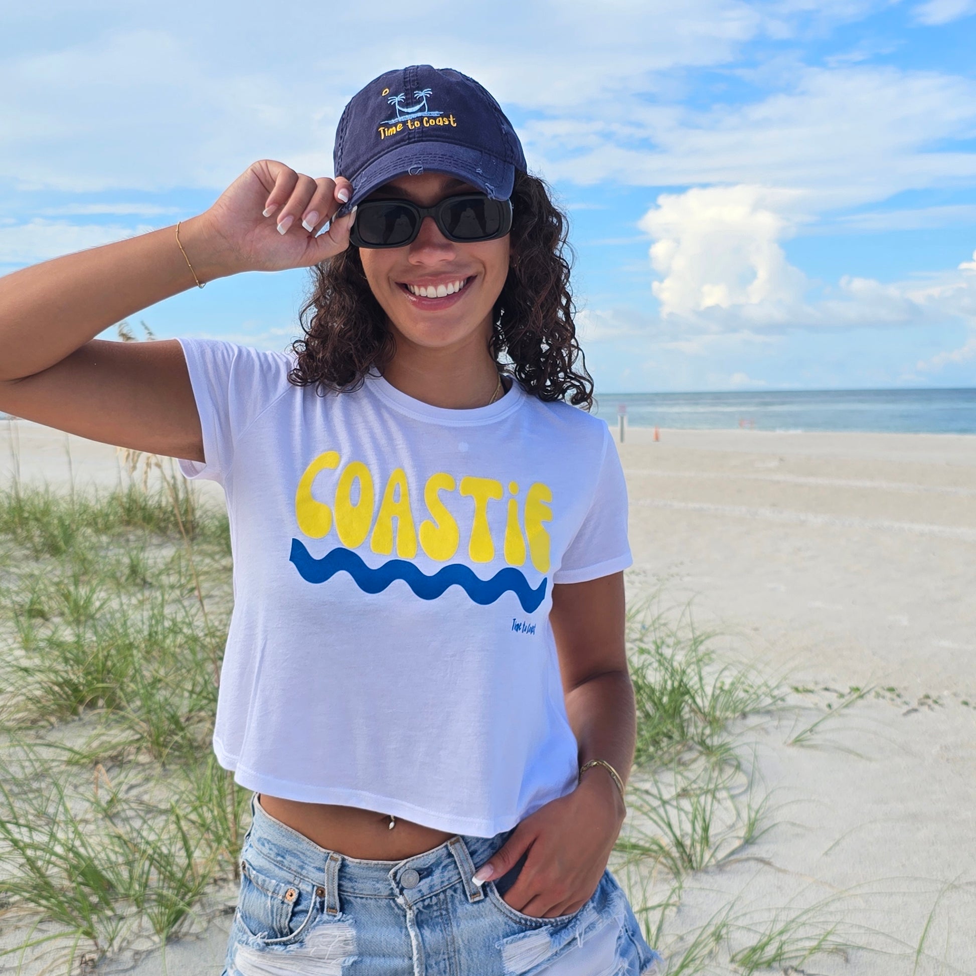 Woman in the sand dunes of the beach, wearing sunglasses, denim shorts, Time to Coast Dad hat and a white Time to Coast Palm Cloth Coastie crop Tee. Coastie graphic is yellow with a blue wave. 