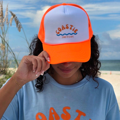 Woman wearing a foam high profile trucker hat.  Graphic is printed with Time to Coast Coastie with waves. Hat color is bright orange bill and mesh back with white front. 