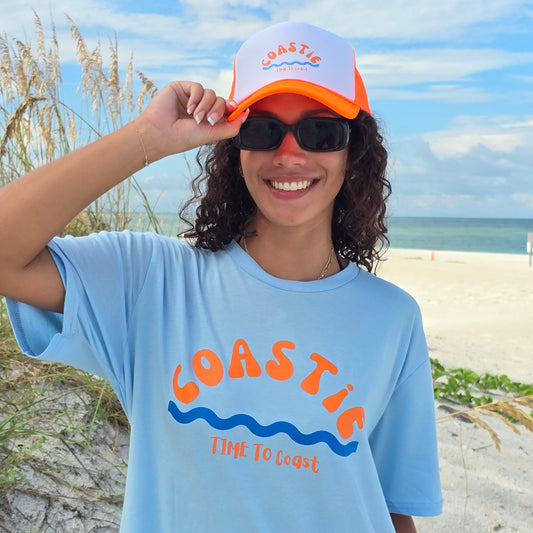 Woman wearing sunglasses, a coastie homegrown tee and a foam high profile trucker hat.  Graphic is printed with Time to Coast Coastie with waves. Hat color is bright orange bill and mesh back with white front. 