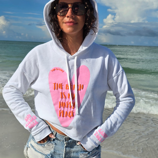 Woman standing on the beach by the ocean wearing sunglasses, denim shorts and a Florida Fleece White Cropped Hoodie Graphic Top, with the hood up.  Graphic is the Time to Coast Heart, Beach is my happy place slogan.  Heart is pink and slogan is red.  Sleeves have pink  words of sandy toes & sun kissed nose. 