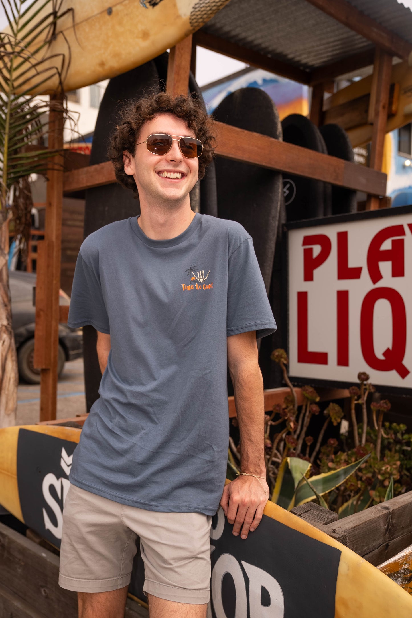 Man wearing sunglasses at a surf shop in a gray blue unisex  graphic  t-shirt in organic cotton made in USA. Homegrown t-shirt shown with left chest graphic with white shaka and  Time to Coast Logo.  
