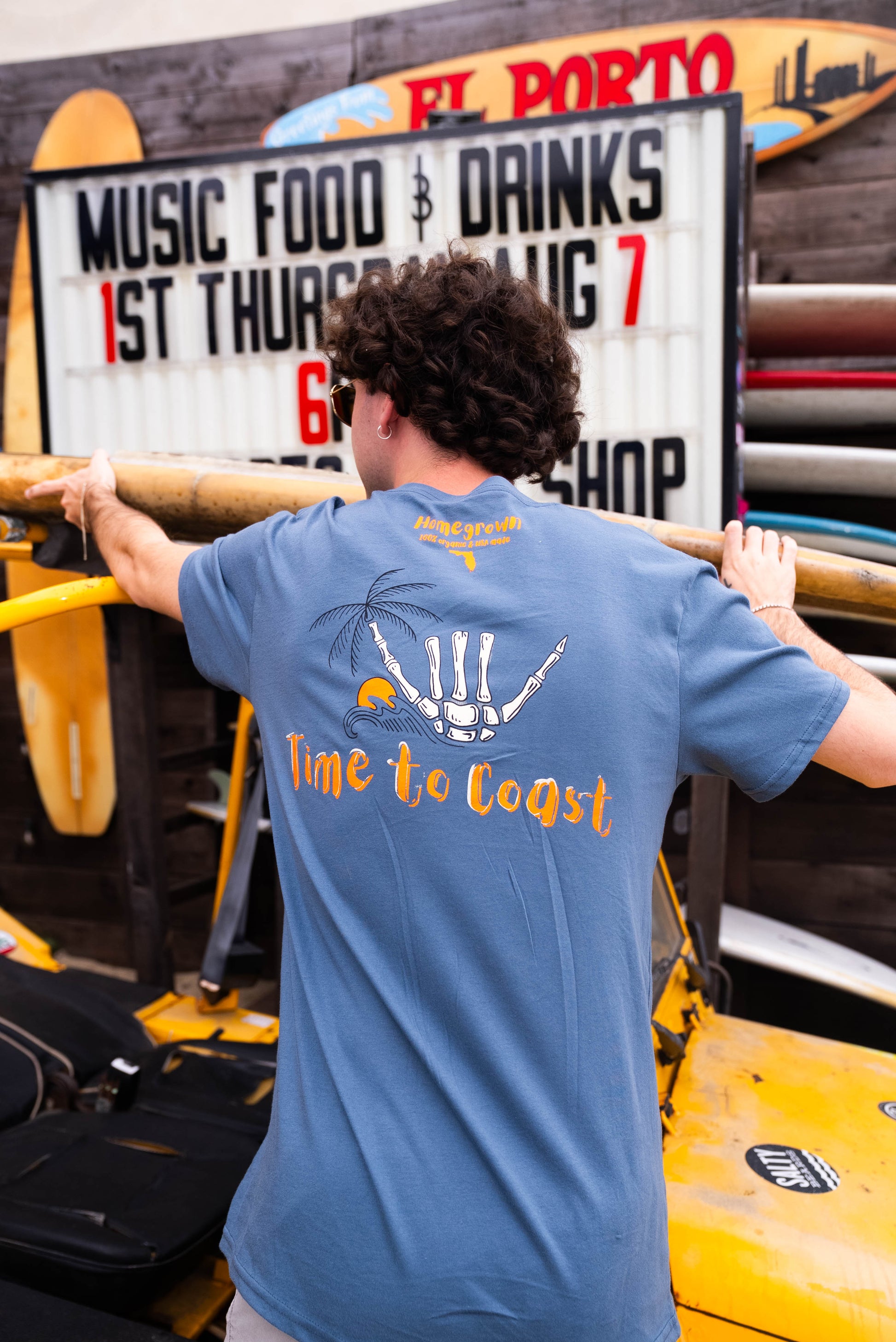 man at a surf shop showing the back of a gray blue color unisex  graphic  t-shirt in organic cotton made in USA. Homegrown t-shirt with is shown with the large back graphic of a shaka hand in white with orange Time to Coast Logo. 