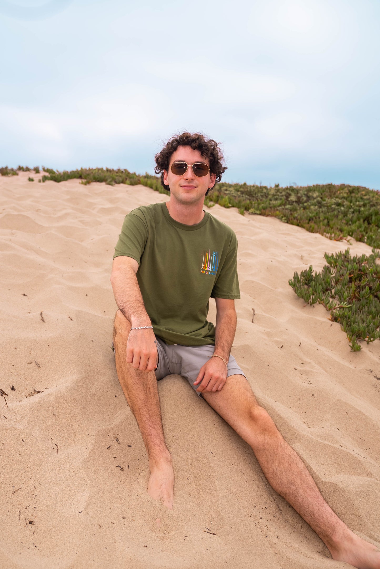 man sitting on the sandy dunes wearing sunglasses, shorts a moss color unisex  graphic  t-shirt in organic cotton made in USA. Homegrown t-shirt shown with left chest graphic multi color surf and sup boards.  Large  back graphic  with same graphic and Time to Coast graphic. 