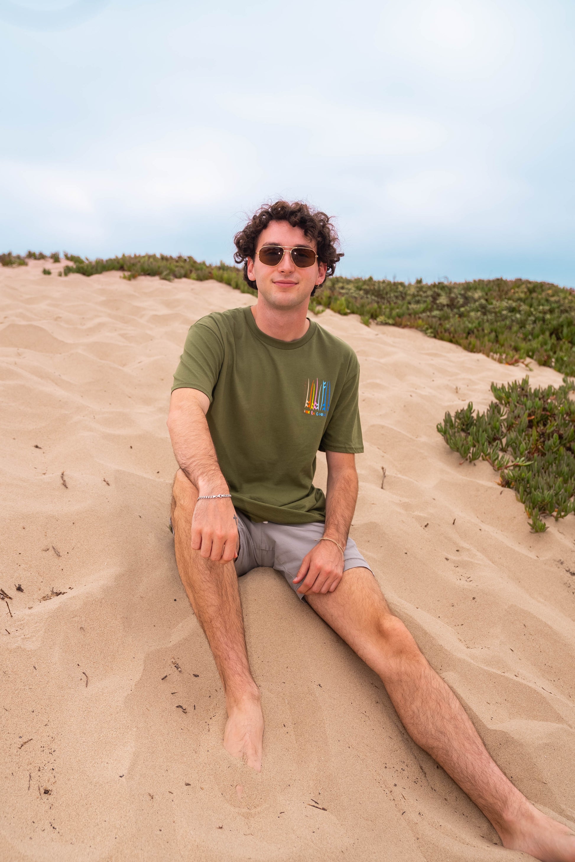 man sitting on the sandy dunes wearing sunglasses, shorts a moss color unisex  graphic  t-shirt in organic cotton made in USA. Homegrown t-shirt shown with left chest graphic multi color surf and sup boards.  Large  back graphic  with same graphic and Time to Coast graphic. 