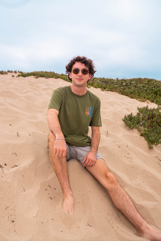 man sitting on the sandy dunes wearing sunglasses, shorts a moss color unisex  graphic  t-shirt in organic cotton made in USA. Homegrown t-shirt shown with left chest graphic multi color surf and sup boards.  Large  back graphic  with same graphic and Time to Coast graphic. 