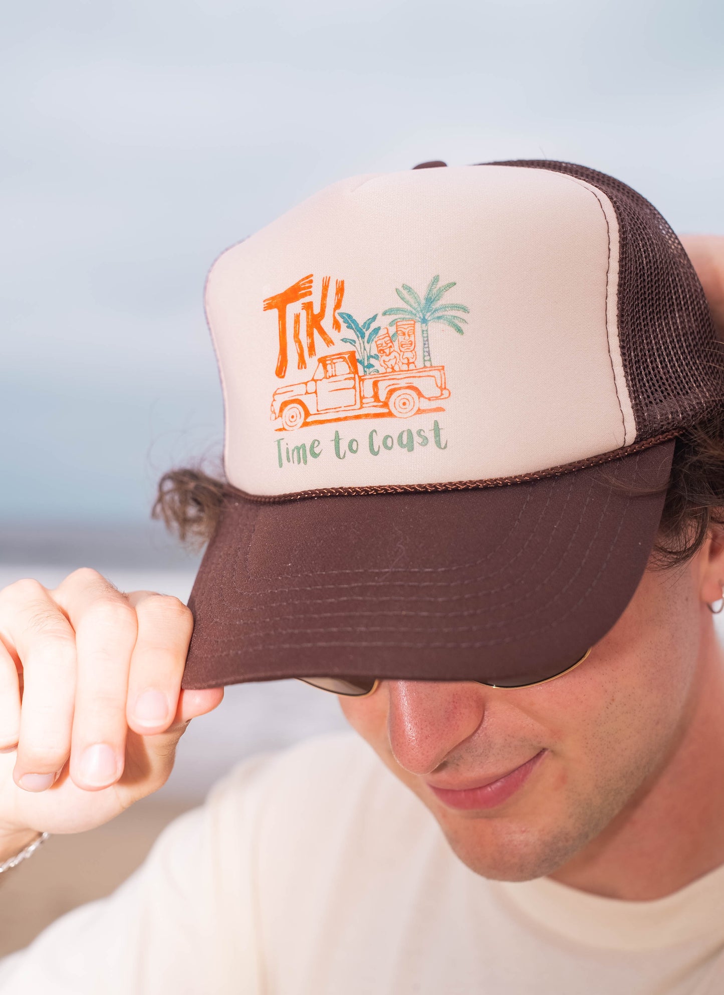 Man wearing a foam high profile trucker hat.  Graphic is printed with Time to Coast Tiki Truck. Hat color is beige and brown with a brown mesh back.