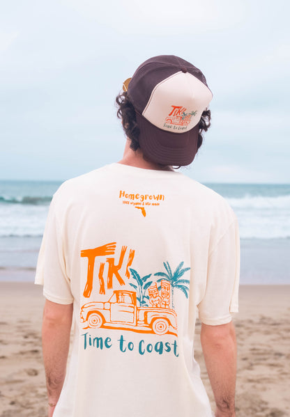 man on the beach wearing a tan and brown truck hat with a tiki truck and showing the back of a creme colored unisex  graphic  t-shirt in organic cotton made in USA. Homegrown t-shirt shown with colorful back graphic of a Tiki Truck and Time to Coast Logo