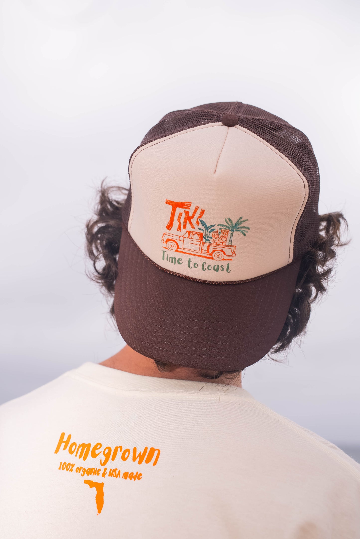 Man wearing a foam high profile trucker hat backwards.  Graphic is printed with Time to Coast Tiki Truck. Hat color is beige and brown with a brown mesh back.