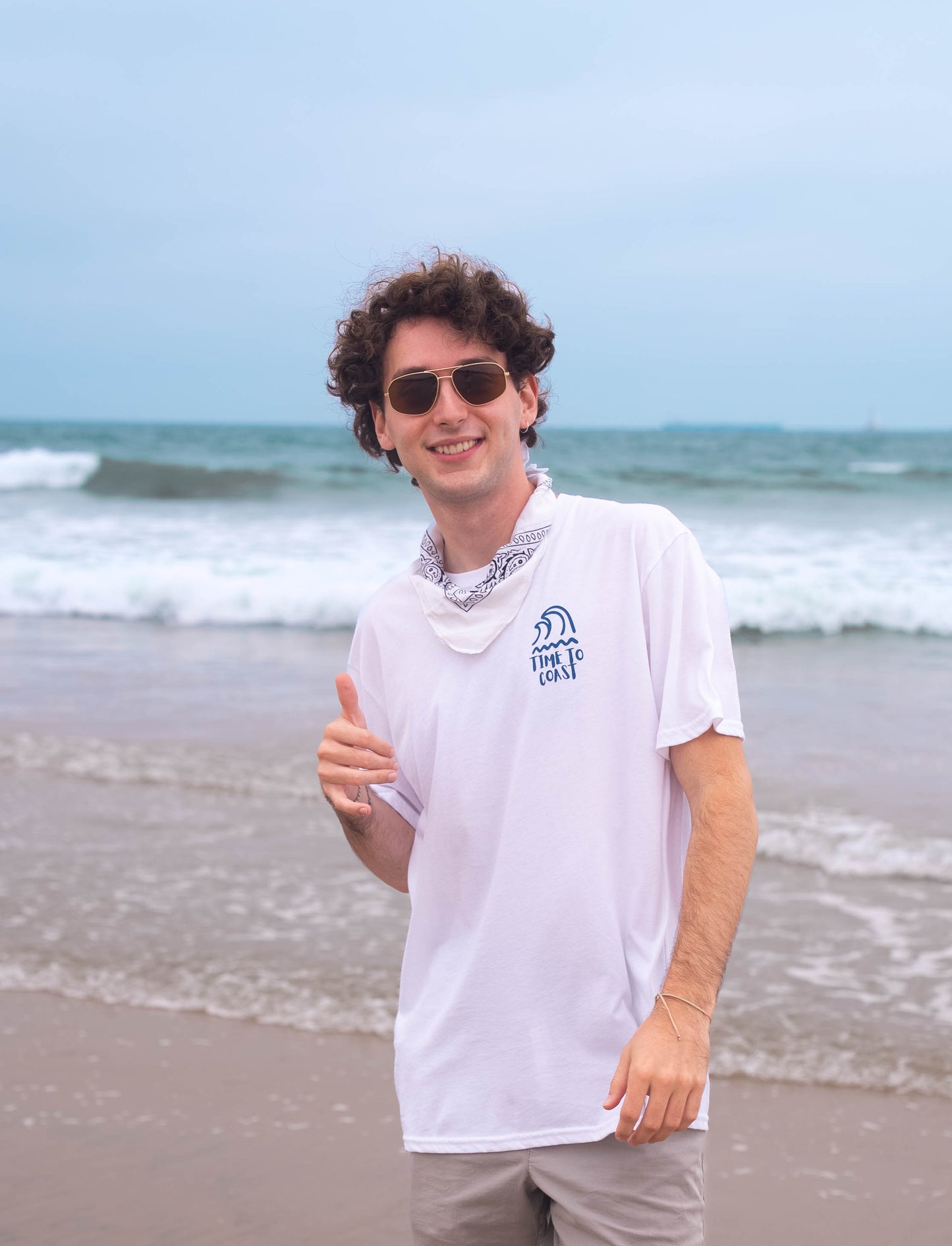 man standing by the ocean with sunglasses and bandana, on the beach wearing a white color unisex  graphic  t-shirt in organic cotton made in USA. Homegrown t-shirt showing the small left chest graphic of swell surf waves.  Also has large back graphic of surf waves and time to coast logo.
