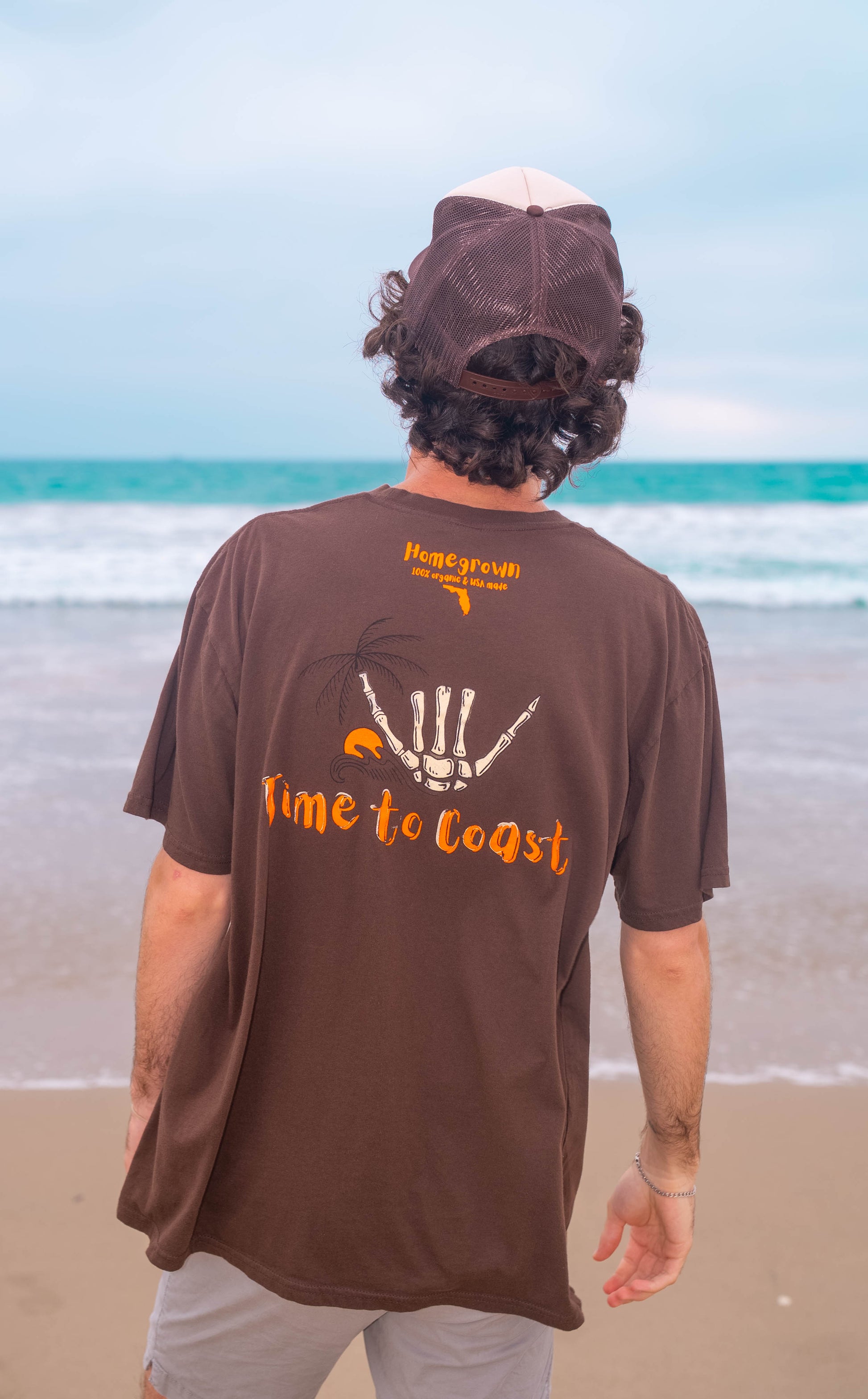 man on the beach showing the back of a bark or brown color unisex  graphic  t-shirt in organic cotton made in USA. Homegrown t-shirt with is shown with the large back graphic of a shaka hand in white with orange Time to Coast Logo. 