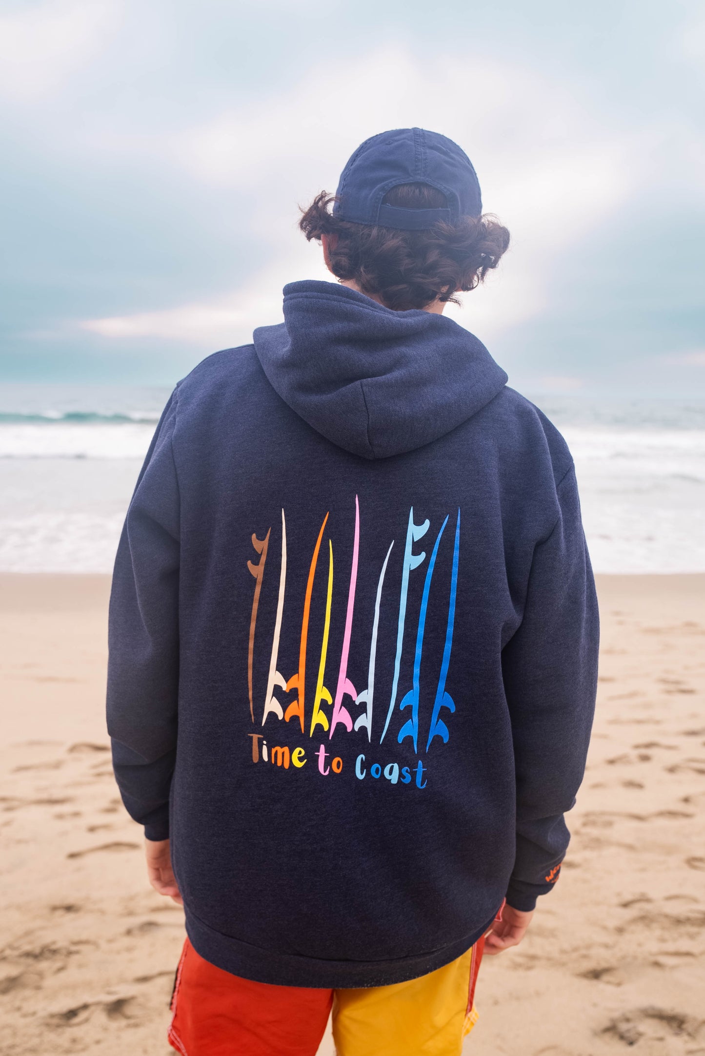 Man on a beach looking out to the ocean, showing the back of the blue indigo Florida Fleece Zip Front Hoodie.  The graphic is the board meeting surf and sup boards in multi colors. Large graphic on back with small left chest graphic.  One sleeve has the words where summer never ends.