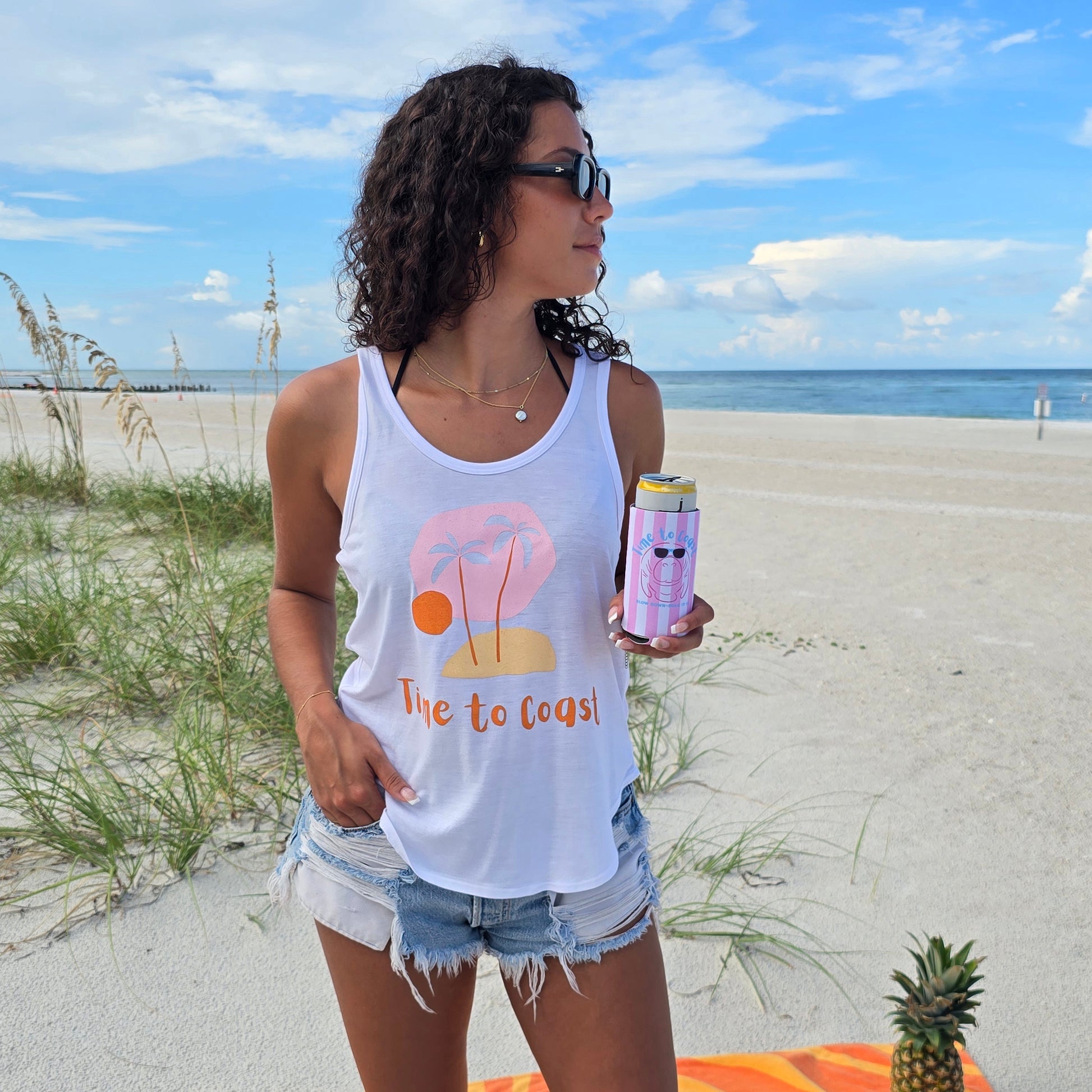 woman wearing a time to coast ,my island palm cloth tank, holding a neoprene slim coozie with a graphic of a manatee wearing sunglasses