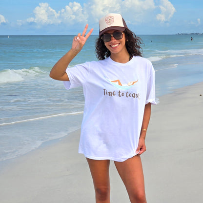 woman on the beach shore wearing a white color graphic  t-shirt in organic cotton made in USA and a tan trucker hat.  Homegrown t-shirt with a large front chest graphic of a woman in a hammock just chilling.  Model is wearing a boyfriend fit size.