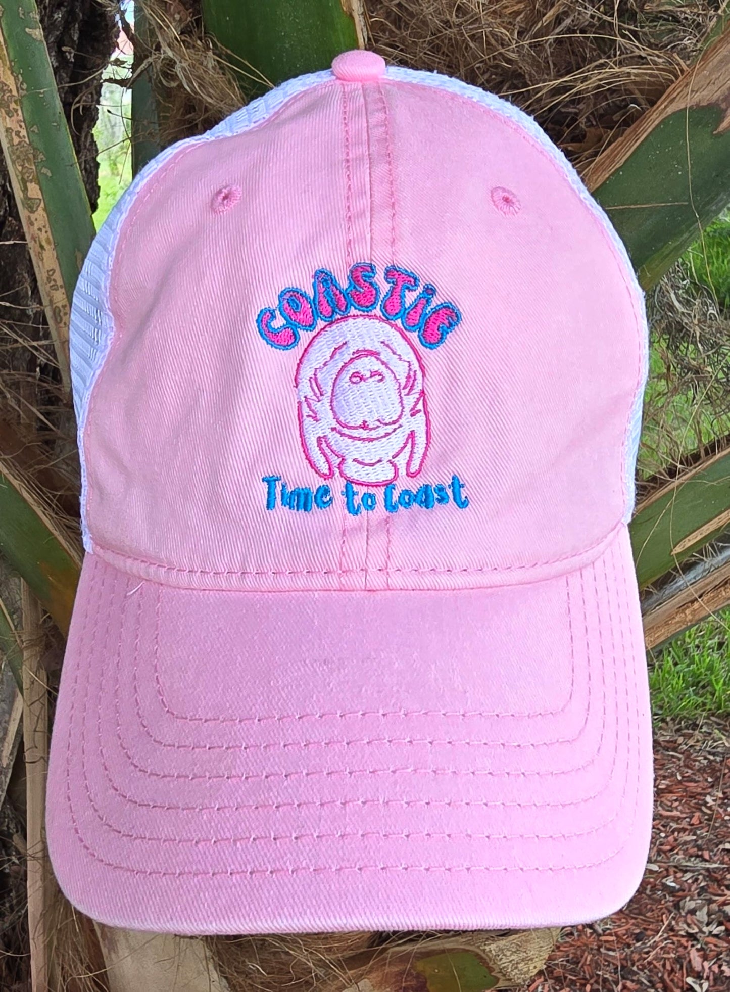 Laydown of the Coastie Manatee embroidered twill low profile trucker hat. Color of hat is washed pink twill with a white embroidered manatee and tan mesh back.