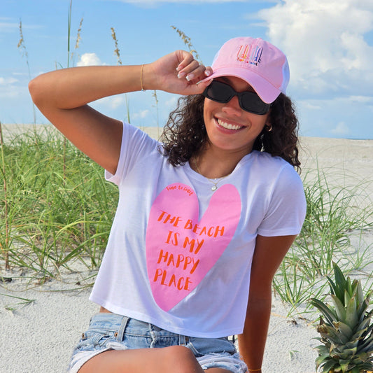 Woman sitting in the sand dunes of the beach, wearing sunglasses, denim shorts, Time to Coast Board Meeting hat and a white Time to Coast Palm Cloth crop tee. Graphic is a heart with beach is my happy place slogan. Heart color is pink with red wording. 