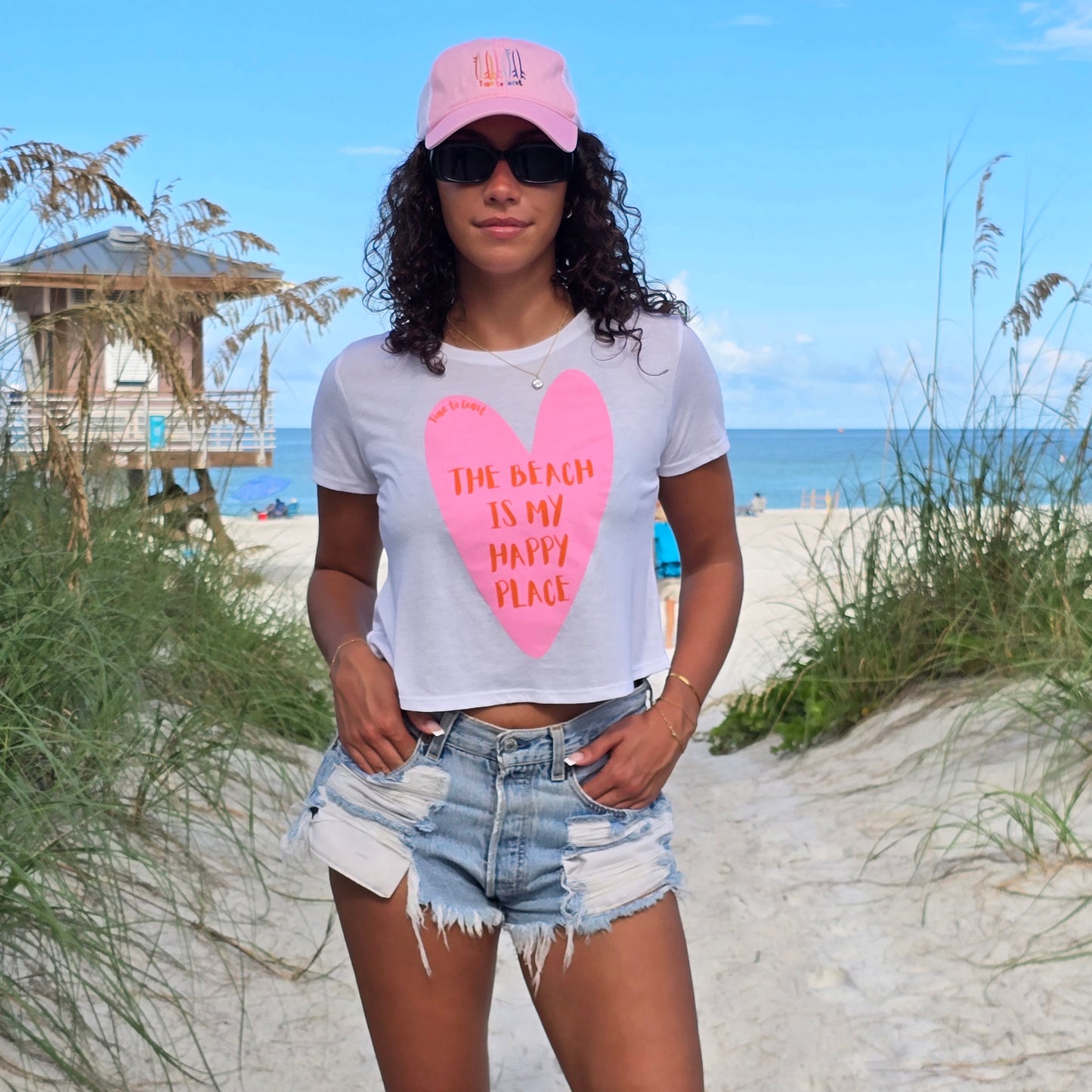 Woman in the sand dunes of the beach, wearing sunglasses, denim shorts, Time to Coast Board Meeting hat and a white Time to Coast Palm Cloth crop tee. Graphic is a heart with beach is my happy place slogan. Heart color is pink with red wording. 