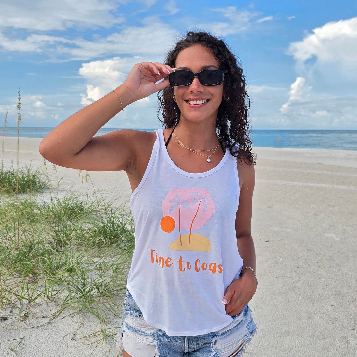 Woman in the sand dunes of the beach, wearing sunglasses, denim shorts and a white Time to Coast Palm Cloth My little island graphic tank top. Graphic is soft colors of pink, yellow and orange.
