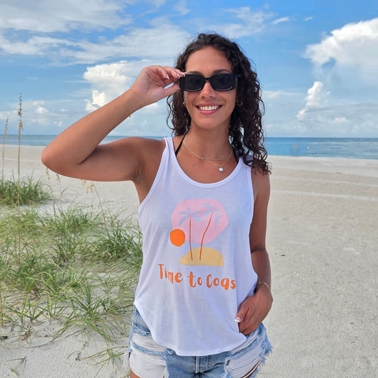 Woman in the sand dunes of the beach, wearing sunglasses, denim shorts and a white Time to Coast Palm Cloth My little island graphic tank top. Graphic is soft colors of pink, yellow and orange.