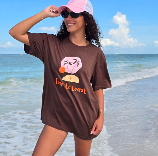 woman on the beach shore wearing a pink trucker hat and a bark or brown colored unisex  graphic  t-shirt in organic cotton made in USA. Homegrown t-shirt with a large front colorful graphic of a small island, two palm trees, sun. wearing a boyfriend fit size. 
