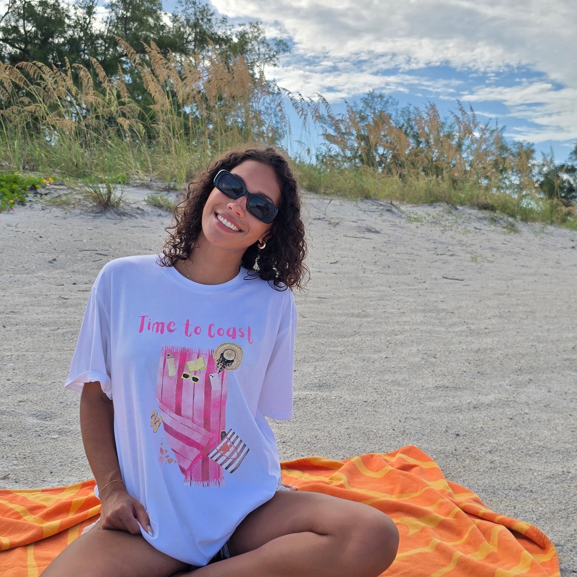 woman sitting on the beach wearing a white color graphic  t-shirt in organic cotton made in USA and a pair of destressed short jean shorts. Homegrown t-shirt with large front colorful graphic of beach towel and beach essentials; hats, sunglasses, water bottle, bag and Time to Coast Logo.  Model is wearing an oversize fit