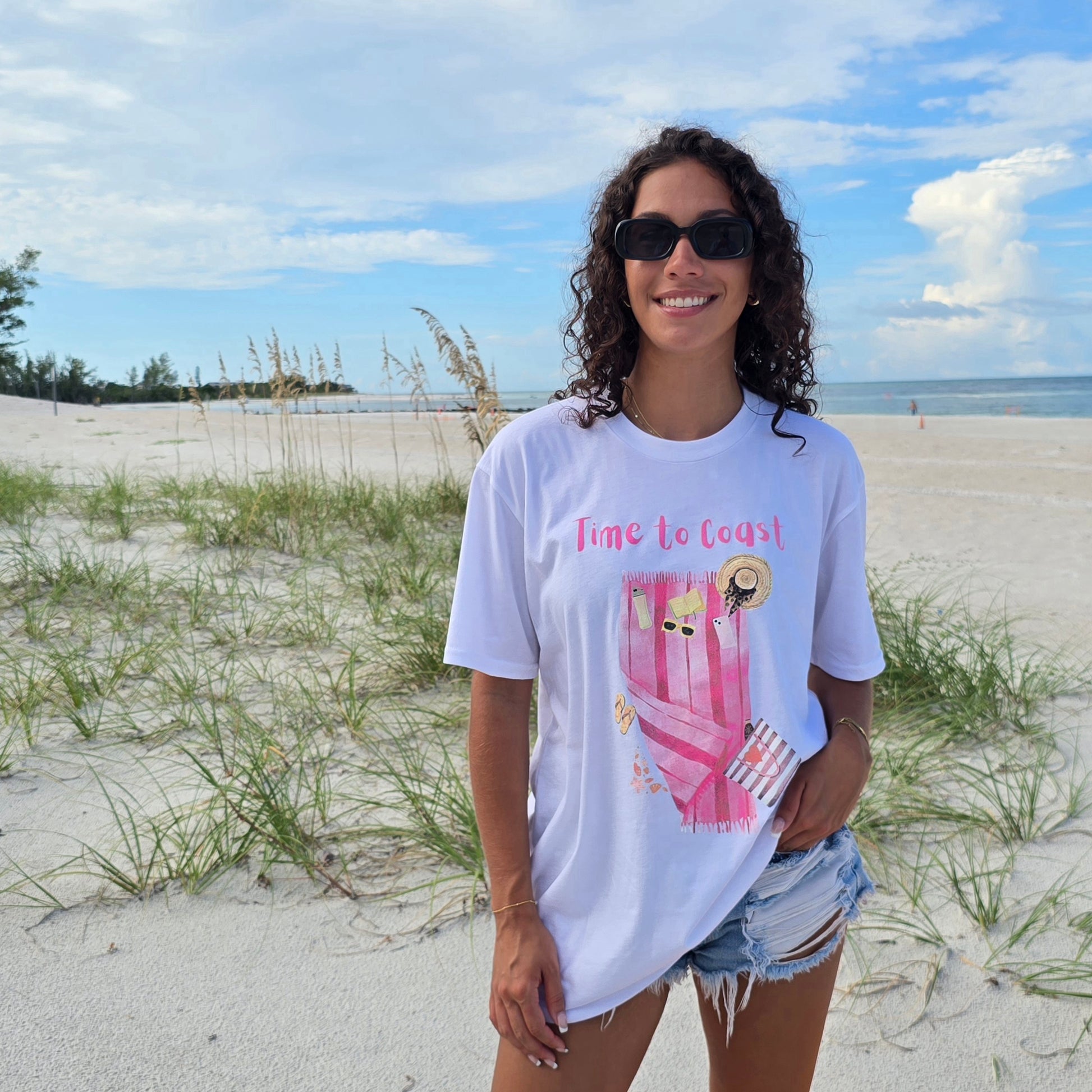woman on the beach wearing a white  color graphic  t-shirt in organic cotton made in USA and a pair of destressed short jean shorts. Homegrown t-shirt with large front colorful graphic of beach towel and beach essentials; hats, sunglasses, water bottle, bag and Time to Coast Logo.  Model is wearing an oversize fit